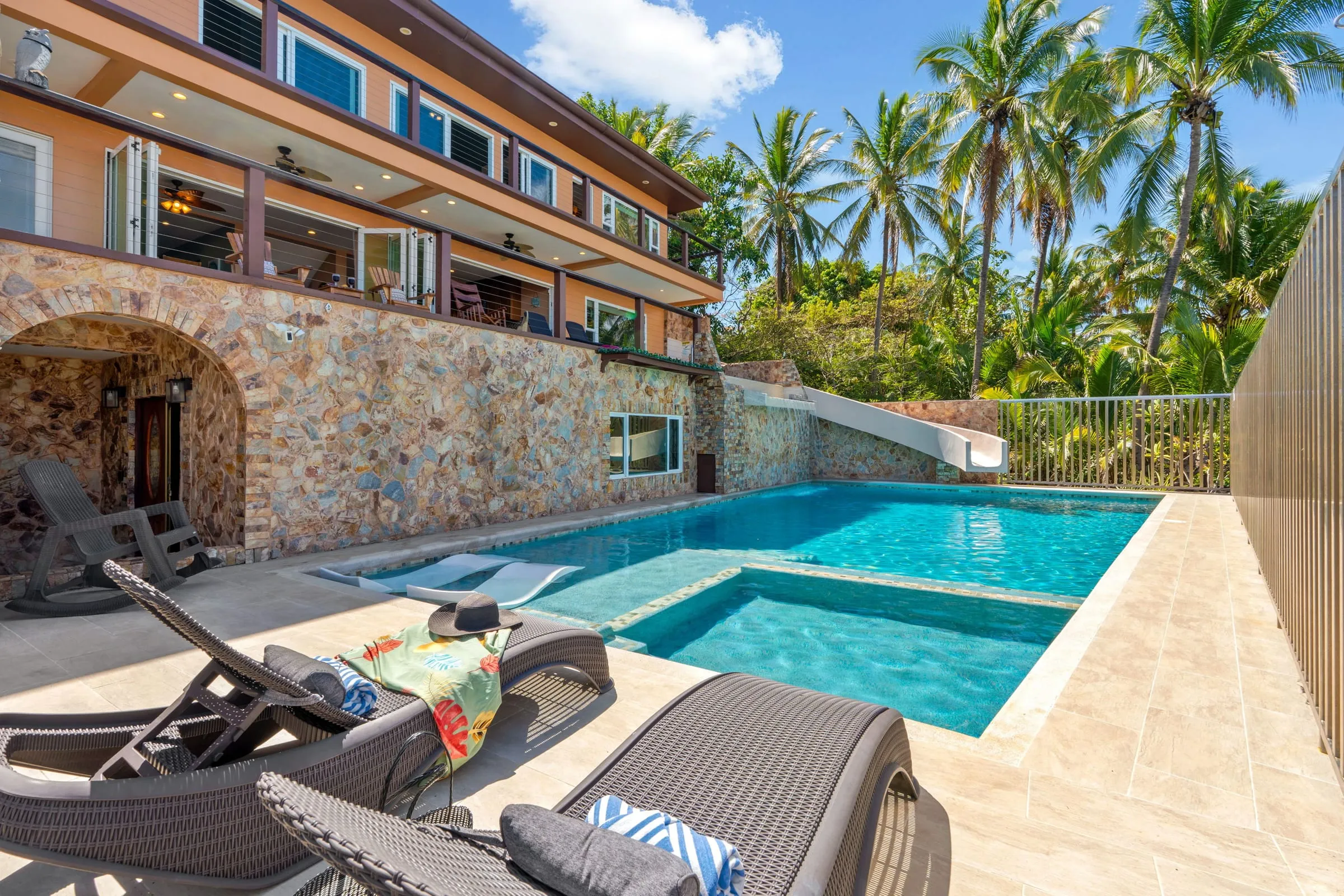 Pool deck with wicker loungers beside the pool and white waterslide, below the stone-walled house and palm trees