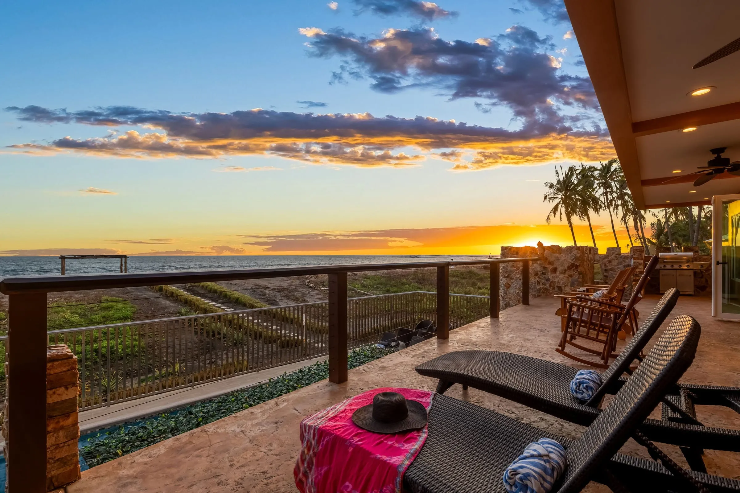 Terraza techada con camastros y mecedoras frente al atardecer sobre el mar
